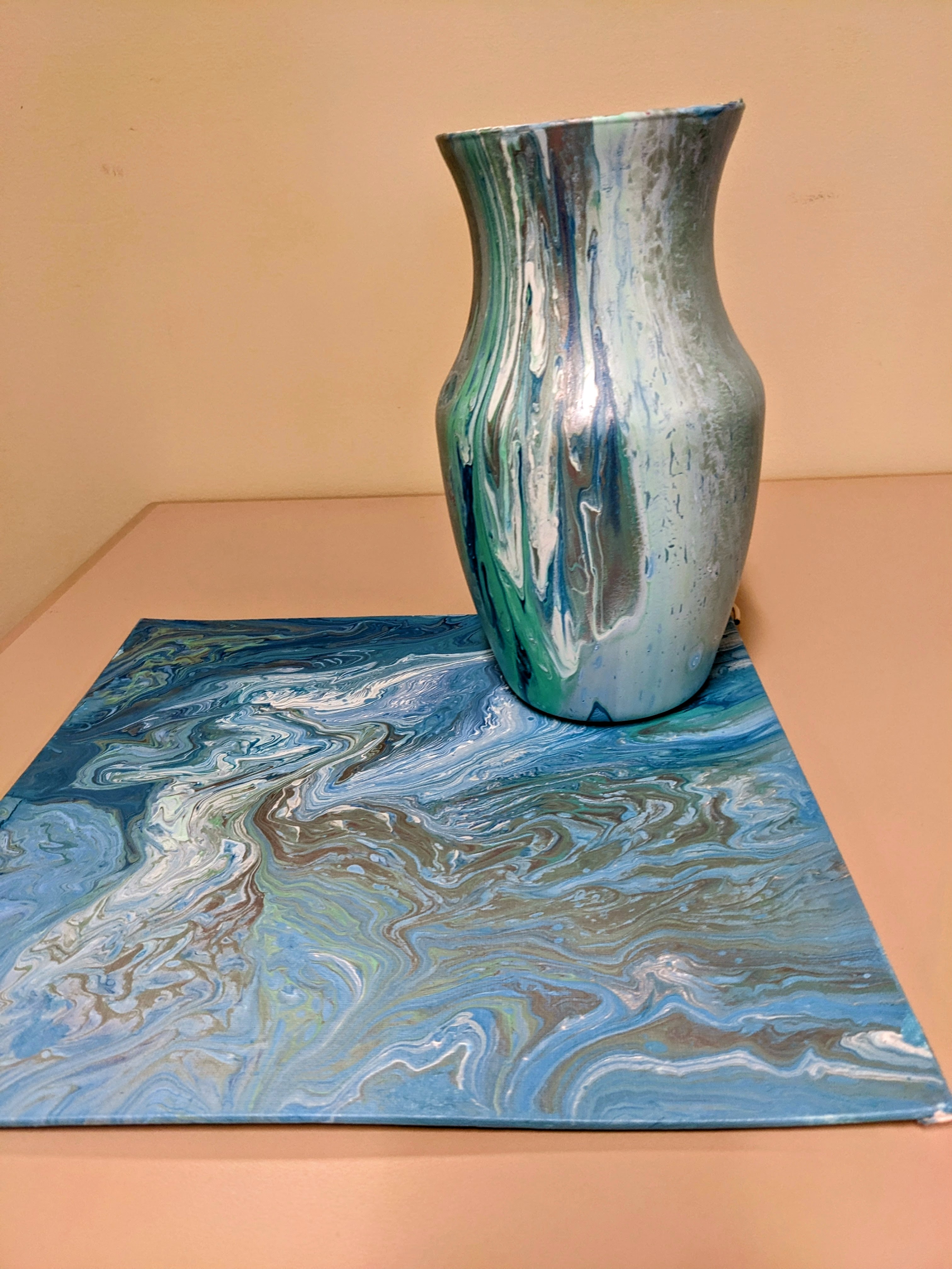 Glass vase and canvas with blue, aqua, silver, and white paint poured over them.