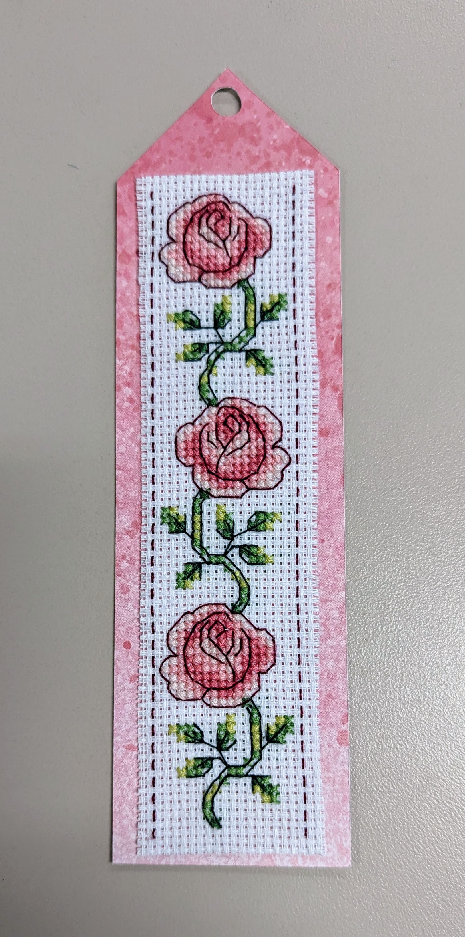 Cross stitch bookmark with 3 pink roses, leaves, & vines on a pink cardstock background.